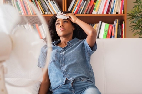 Person cooling off with fan, sitting on couch, surrounded by books.