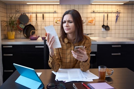 A woman in a kitchen reviews bills with a tablet and smartphone.
