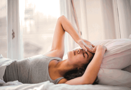 Woman waking up in bed, stretching and shielding eyes from morning light.