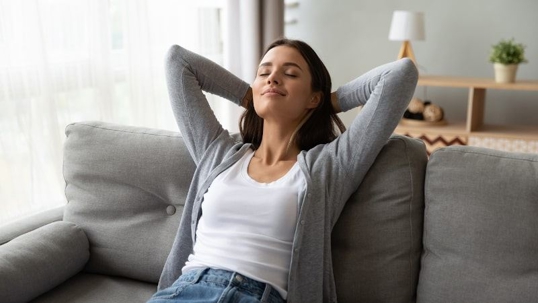 A woman relaxing on a couch with eyes closed and arms behind her head.